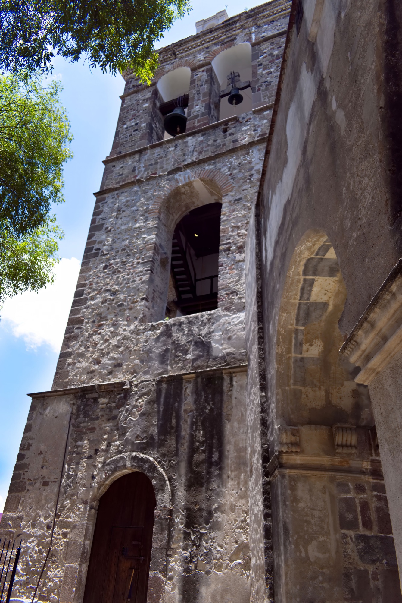 A historic stone bell tower and archway framed by leafy trees and blue sky