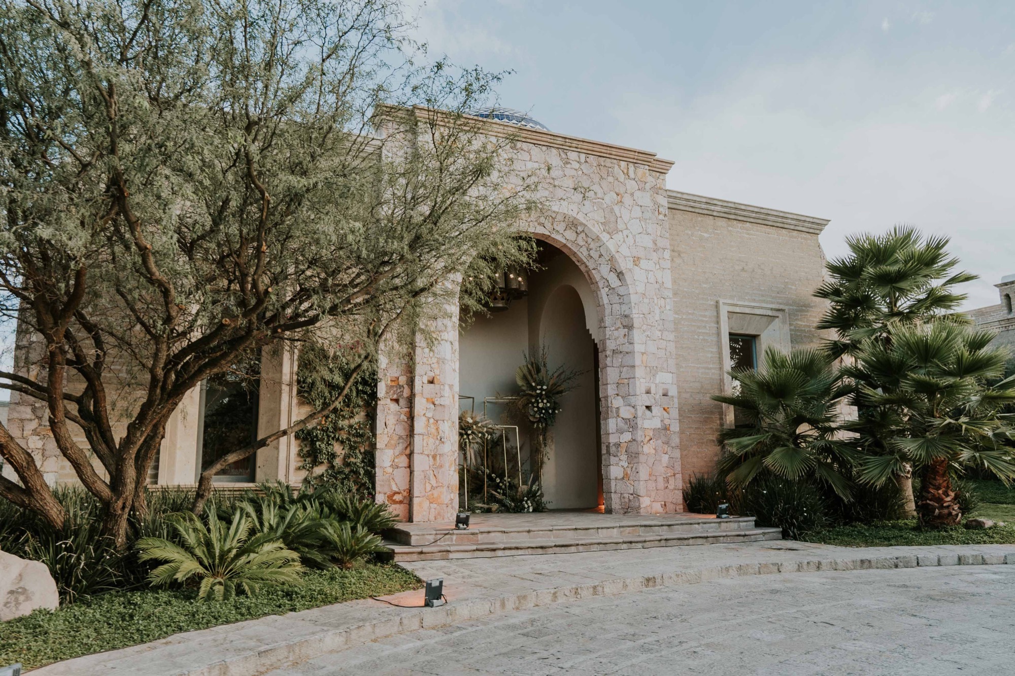 A pale stone building with a welcoming arch surrounded by palms and native greenery